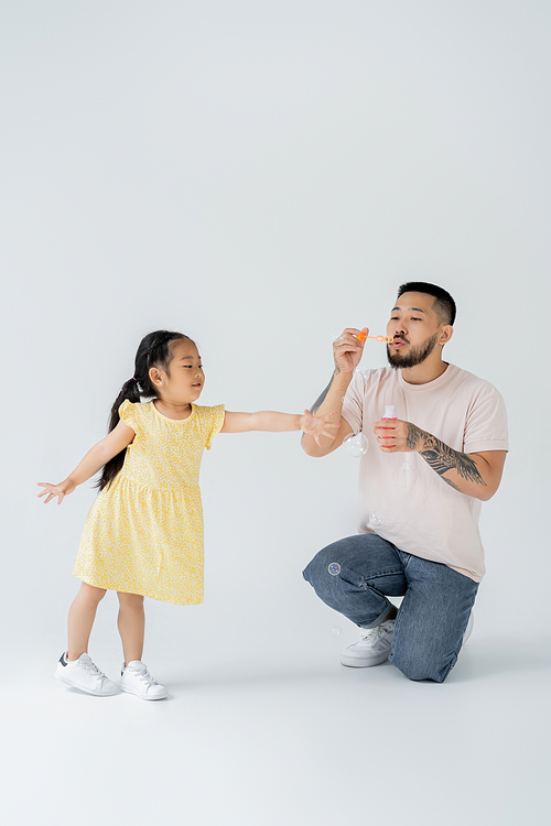 asian father blowing soap bubbles near cheerful preschooler daughter on grey