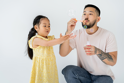asian father blowing soap bubbles near amazed preschooler daughter isolated on grey