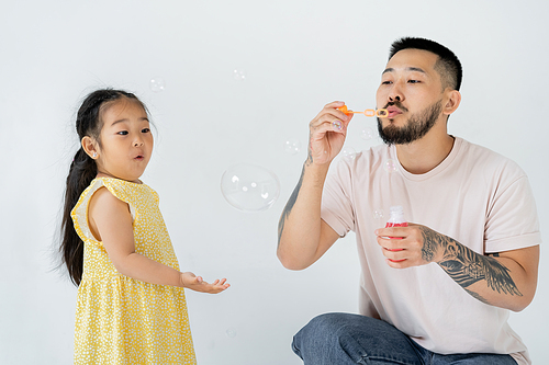 asian father blowing soap bubbles near surprised preschooler daughter isolated on grey