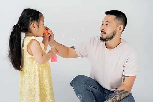 asian preschooler kid in yellow dress blowing soap bubbles near tattooed father on grey