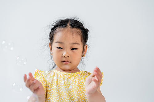 portrait of asian preschooler girl in yellow dress near blurred soap bubbles isolated on grey