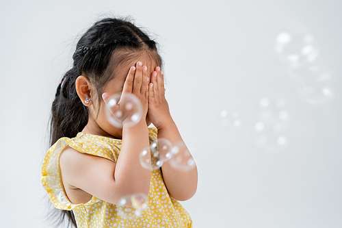 portrait of asian preschooler girl in yellow dress covering face with hands near soap bubbles isolated on grey