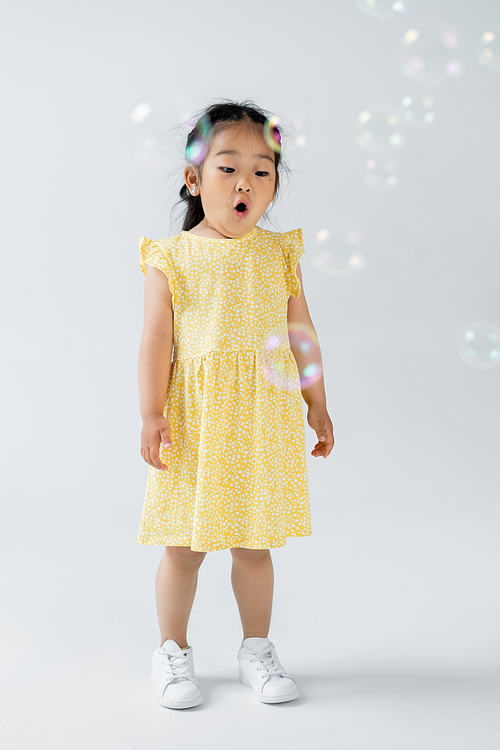 full length of shocked asian preschooler girl in yellow dress standing soap bubbles on grey