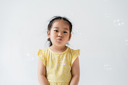 portrait of asian preschooler girl in yellow dress pouting lips near near soap bubbles isolated on grey