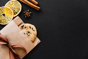 Top view of cookie in craft package, dry orange slices and spices on black background