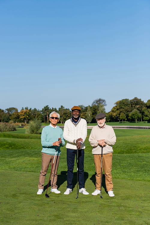 asian man in sunglasses standing near senior multiethnic friends in flat caps with golf clubs