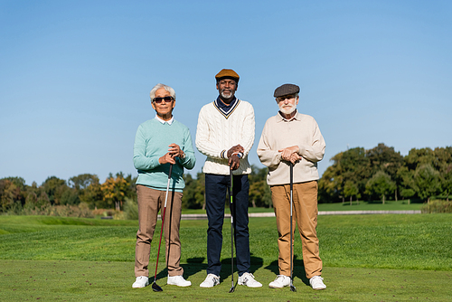 senior asian man in sunglasses standing near multiethnic friends with golf clubs