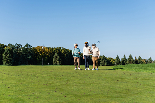 senior asian man in sunglasses walking near multiethnic friends with golf clubs