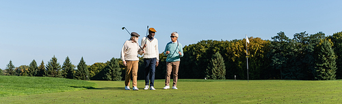 senior interracial friends walking with golf clubs on green field, banner