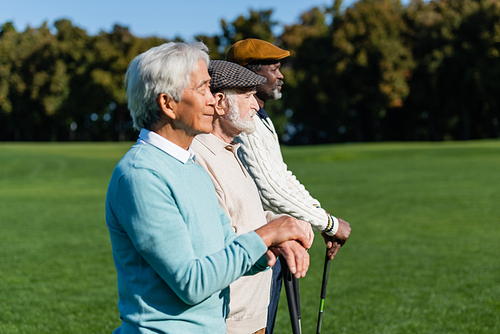 side view of senior man in flat cap near interracial friends with golf clubs