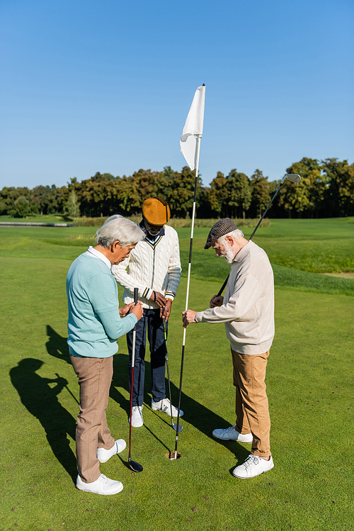 senior man in flat cap holding flag stick near multiethnic friends on golf field