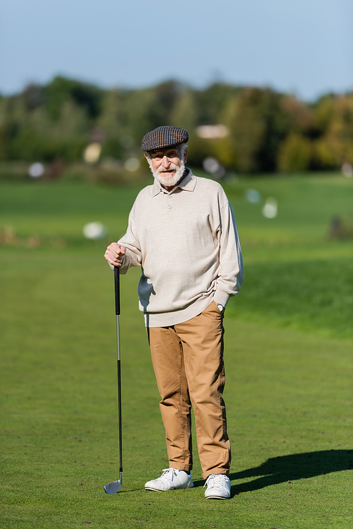 senior man in flat cap standing with hand in pocket and golf club on green field