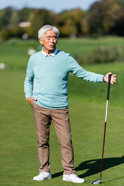 asian senior man standing with hand in pocket and golf club on green field