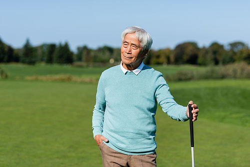 asian senior man standing with hand in pocket and holding golf club on green field