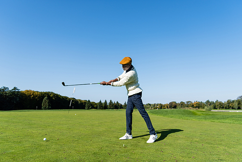 full length of african american man in flat cap holding golf club and playing on green lawn