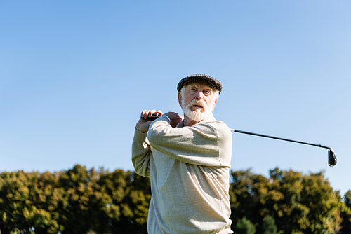 low angle view of bearded senior man in flat cap holding golf club while playing outdoors