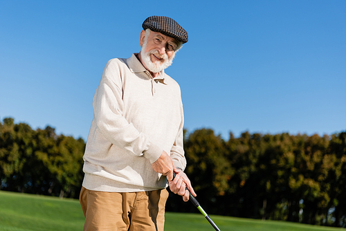 happy senior man in flat cap holding golf club while playing outdoors