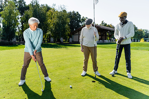 senior man in flat cap holding flag stick near hole and multiethnic friends