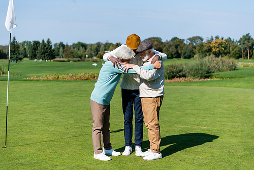 senior multiethnic men hugging near flag stick on green field