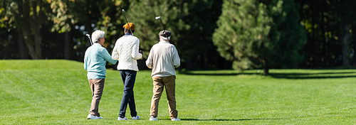 back view of senior multiethnic friends walking with golf clubs on green field near trees, banner