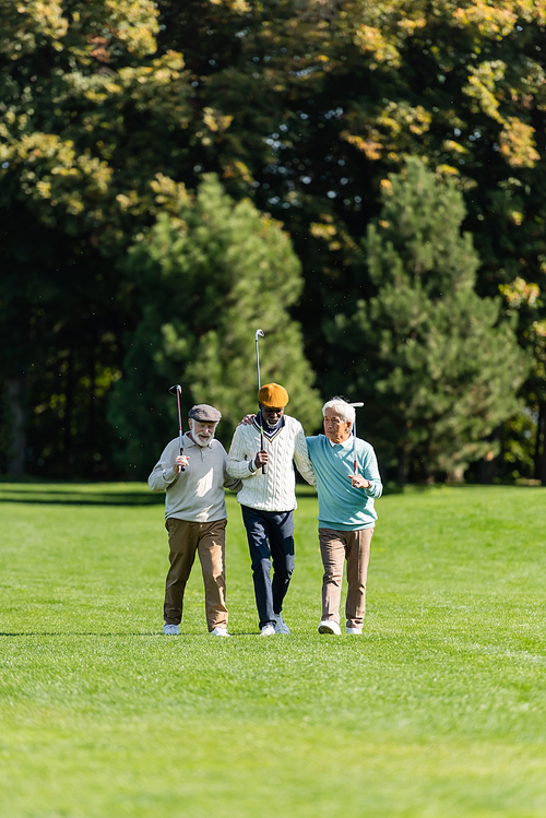 senior multiethnic friends walking with golf clubs on green field near trees