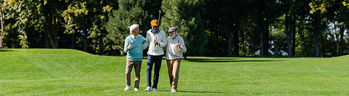 senior multiethnic men walking with golf clubs on green field near trees, banner