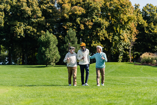 senior multiethnic friends smiling and walking with golf clubs on green field