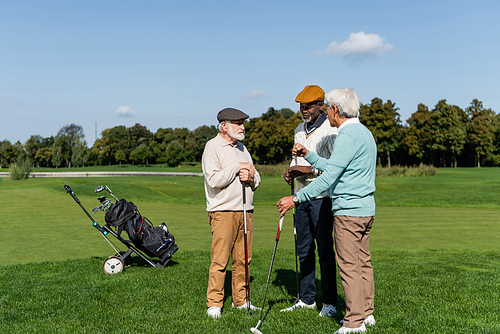 senior multiethnic friends holding golf clubs and talking on field