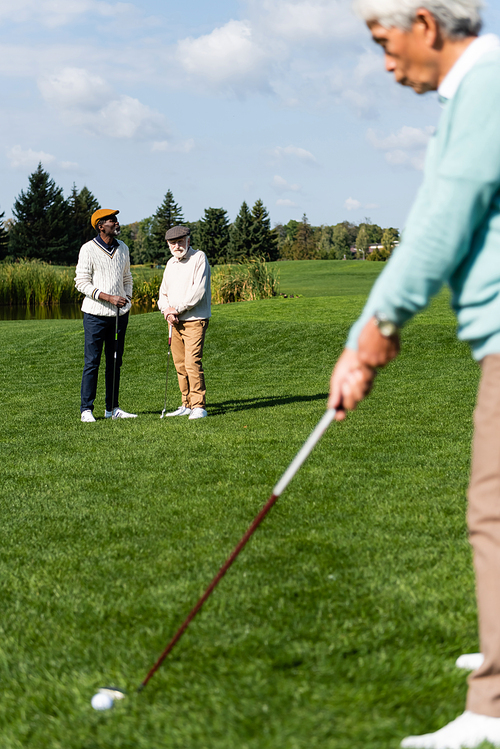 senior interracial men in flat caps looking at asian friend playing golf on blurred foreground