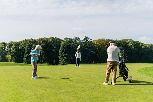 senior asian man playing golf near interracial friends with flag stick and golf cart