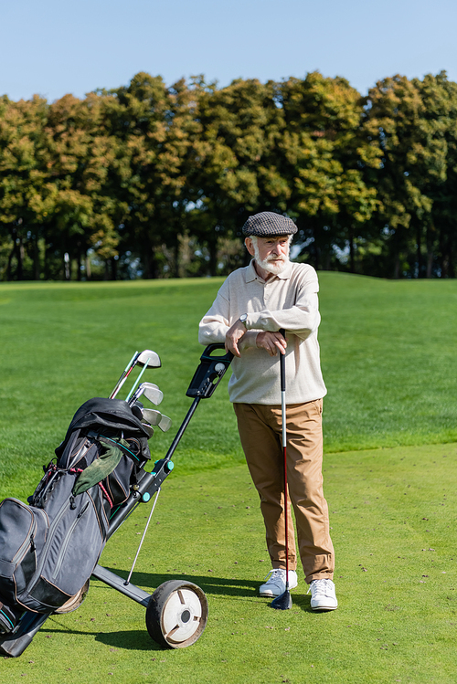 bearded senior man in flat cap standing near golf cart