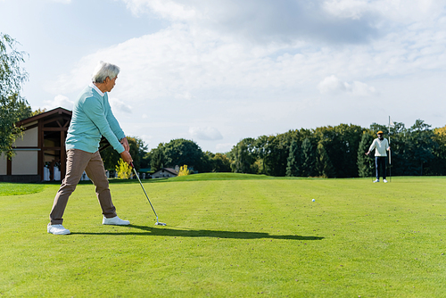 senior asian man playing golf near wealthy african american friend with flag stick