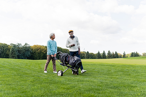 asian senior man walking with golf cart near african american friend