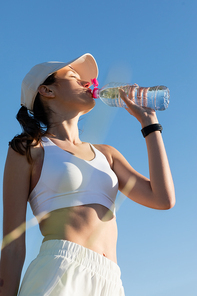 low angle view of sportive woman in  and sports bra drinking refreshing water against blue sky