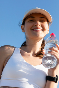 low angle view of smiling woman in  and sports bra holding bottle with water against blue sky