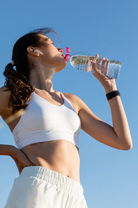 low angle view of young woman in wireless earphone drinking water against blue sky