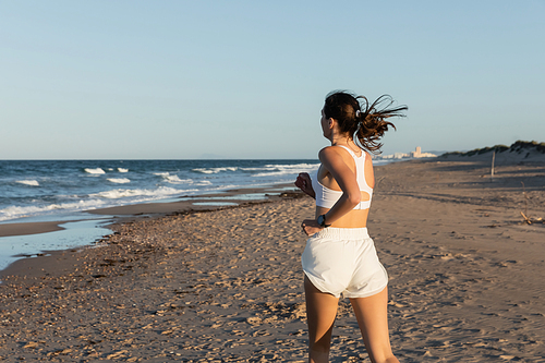 young brunette woman in white sportswear jogging on sandy beach in summer
