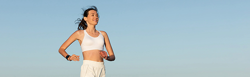 smiling young woman in sportswear jogging against blue sky, banner