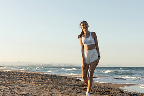 happy woman in white sports bra and shorts standing near sea on beach