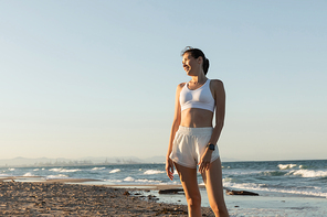cheerful woman in white sports bra and shorts standing near sea on beach