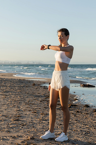 young woman in white sports bra and shorts looking at fitness tracker near sea on beach