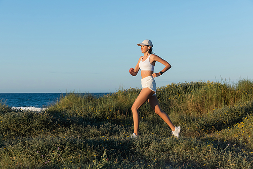 sportive woman in  and wireless earphone running on grass near sea