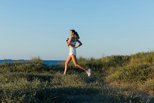young sportive woman in shorts and wireless earphone running on grass near sea