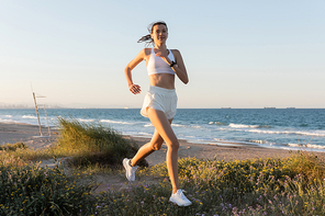 smiling young woman in shorts and wireless earphones jogging on grass near sea