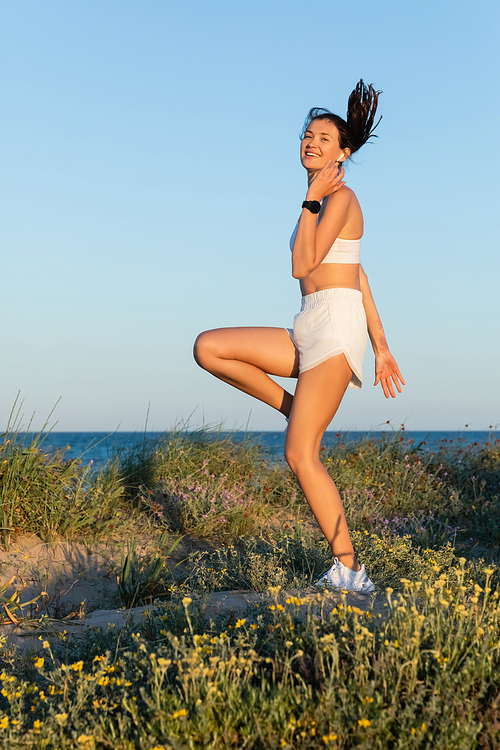 happy fit woman in sportive shorts and wireless earphone jumping near blue sea