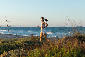 side view of young woman in sports bra and wireless earphone jogging on grass near sea shore