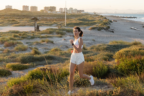 fit woman in shorts and sports bra listening music and running near sea shore
