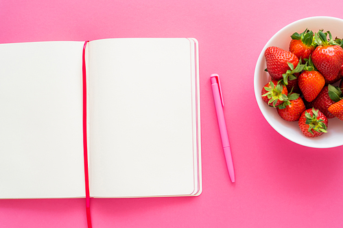 Top view of open notebook with pen and fresh strawberries on pink background