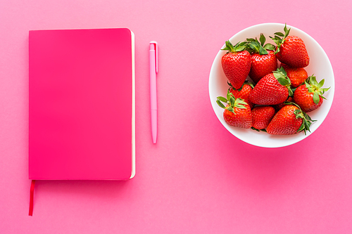 Notebook near pen and whole strawberries in bowl on pink background