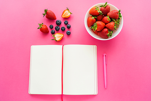 Top view of ripe berries near open notebook on pink background